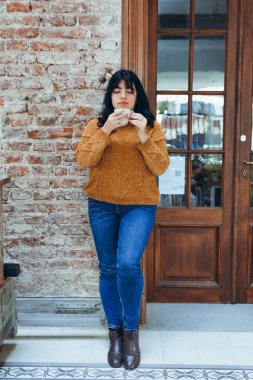 Kahverengi kazaklı ve mavi kot pantolonlu Latin bir kadının dikey görüntüsü. Ahşap kapının yanında duruyor ve kafeteryada kahve içiyor. Boşluğu kopyala.