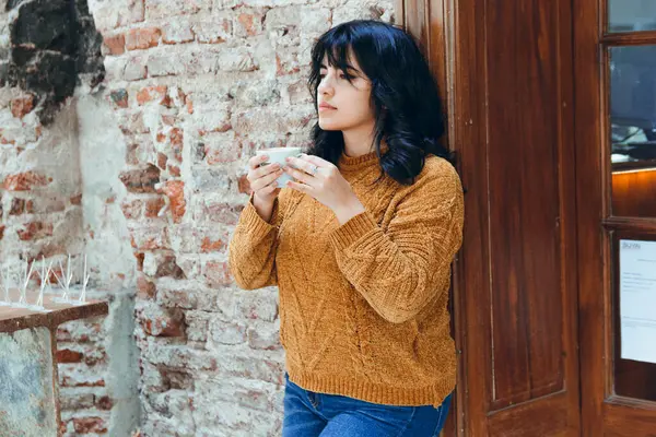 Vertical image of young Latina woman with brown sweater and blue jean pants, she is standing next to wooden door and is drinking coffee in coffee shop. copy space.