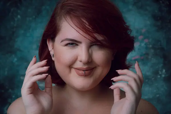 Close-up portrait of plus size young Latina Argentinian woman with short red hair, with earring in her lip and natural makeup, she is smiling and looking at camera happily. with artistic background