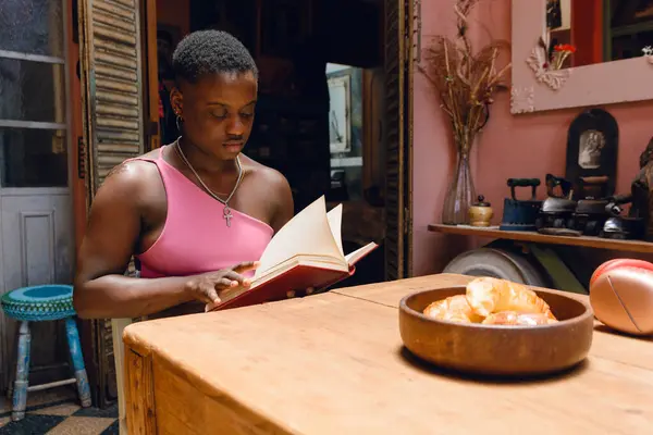young Haitian woman with short hair at home sitting in living room reading comfortably, with croissants on brown ceramic wave plate on table, lifestyle concept, copy space.