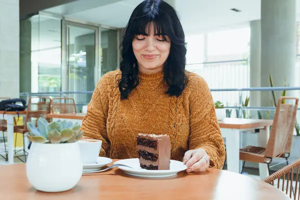 young latin woman with black hair and brown sweater, smiling sitting in cafeteria, happy to eat chocolate cake with coffee, food concept, copy space.
