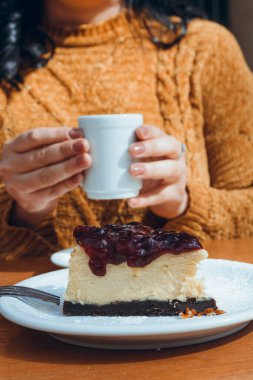 Bir dilim ahududu soslu cheesecake, açık havada servis edilen tabakta, arka planda bulanık kahve fincanı tutan kadın elleri, dikey görüntü, kopyalama alanı