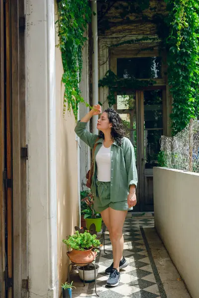 Vertical image of young Latina woman with curls, wearing green clothes, standing at entrance of her house caressing leaf of creeper plant on wall, people and natural conservation concept, copy space.