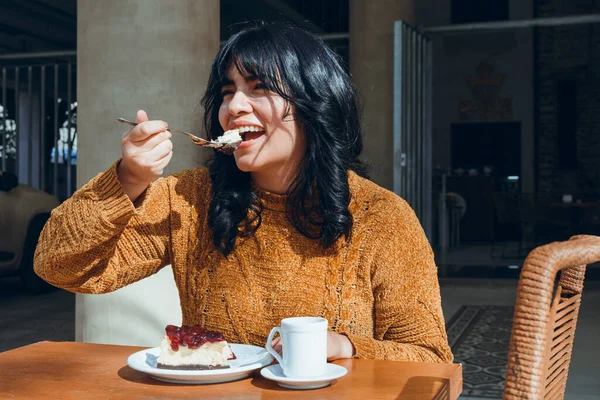 young latin woman with black hair, wearing brown clothes, enjoying afternoon outdoors sitting happily, smiling, with fork eating cake with coffee, coffee shop concept copy space.