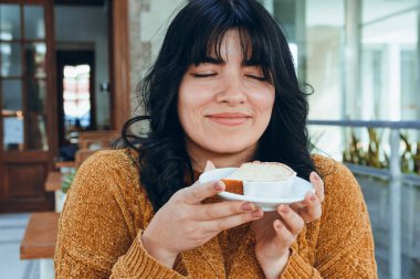 Kafeteryada oturan, uzun saçlı, kahverengi elbiseli, kapali gözlü, genç Latin bir kadin, mutlu bir sekilde yiyecegi puding aromasını hisseder.