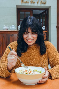 Kahverengi kazaklı, uzun siyah saçlı genç Latin kadın restoranda oturup yemek yiyeceği lezzetli salatayı izlerken mutlu. Restoran konsepti, fotokopi alanı.