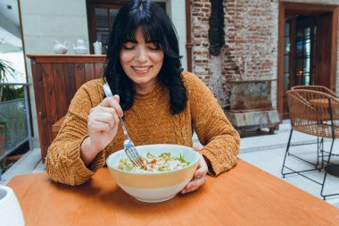 Kahverengi kazaklı, uzun siyah saçlı genç Latin kadın restoranda oturup yemek yiyeceği lezzetli salatayı izlerken mutlu. Restoran konsepti, fotokopi alanı.