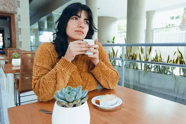 young Latin Venezuelan woman with long black hair, sitting alone in cafeteria drinking coffee and eating pudding alone and thoughtful, relaxed, enjoying moment with herself.