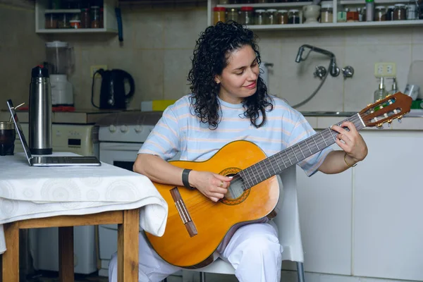 young Venezuelan woman at home sitting happy smiling, with her laptop on table and in online guitar class, rehearsing learning to play new songs. music concept.