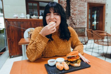 Genç Venezuelalı kadın, lokantada gözleri kapalı ve aç bir şekilde oturuyor ve mutlu bir şekilde fast food, tekeno, hamburger, restoran ve yaşam tarzı konsepti yiyor..