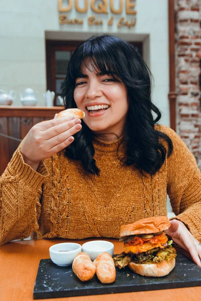 vertical image of young Latina with black hair and brown clothes, happy sitting in restaurant eating tequenos, traditional Venezuelan food, with hamburger and sauce.