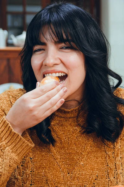 vertical image of young Latina with black hair and brown clothes, happy sitting in restaurant eating tequenos looking at camera, traditional Venezuelan food, with hamburger and sauce.