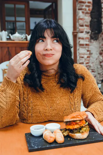 vertical image of young Latina with black hair and brown clothes, happy sitting in restaurant eating tequenos, traditional Venezuelan food, with hamburger and sauce.