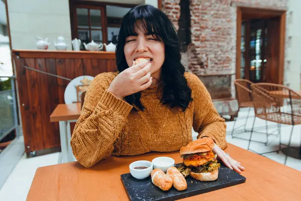 young Venezuelan woman, sitting in restaurant with eyes closed and hungry happy eating fast food, tequenos and hamburger, restaurant and lifestyle concept.