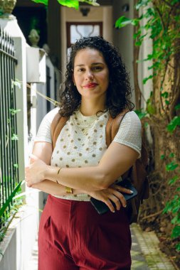 Güçlü genç Latin kadının dikey portresi. Evin girişinde kolları çapraz duruyor. Elinde telefonla gülümsüyor, poz veriyor ve kameraya bakıyor.