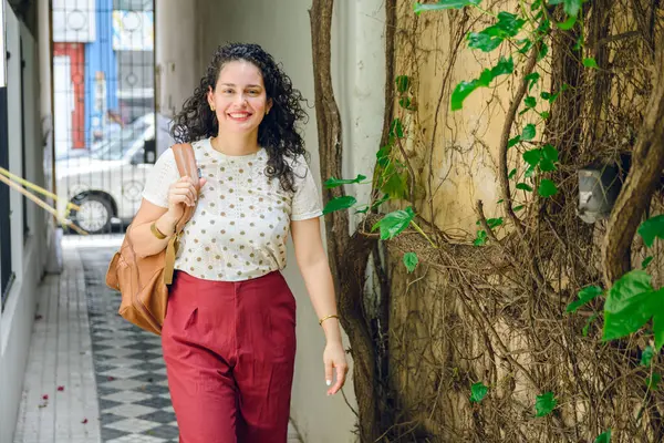 young elegant Venezuelan latina woman, in curlers, with backpack, smiling happy in hallway of residence arriving home, lifestyle concept with copy space.