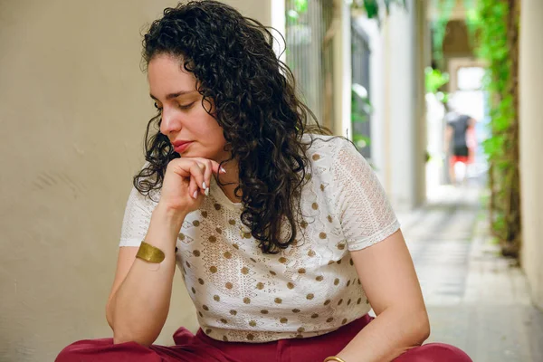 Front view of young Latina woman with curls and Venezuelan ethnicity, she is sitting on floor of entrance with her head down, worried, meditative and thoughtful.