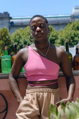 Haitili etnik kökenli genç afro kadının dikey portresi. Kısa saçlı, evde duruyor. Eli cepte kameraya bakıyor.