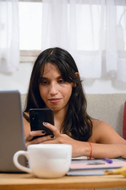 Genç esmer bayan öğrencinin dikey portresi. Evde dikkatle telefonunu izliyor. Masada dizüstü bilgisayarıyla oturuyor..