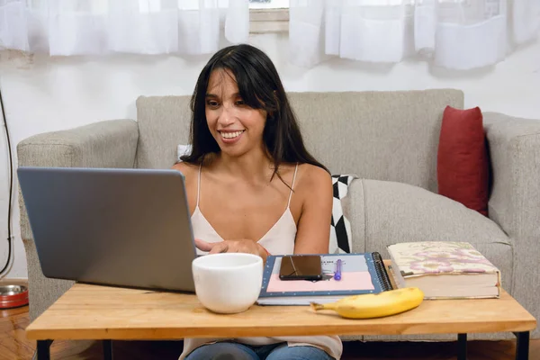 young brunette latin woman happy smiling at home sitting on floor working with her laptop on table in living room, typing on computer, managing her digital business