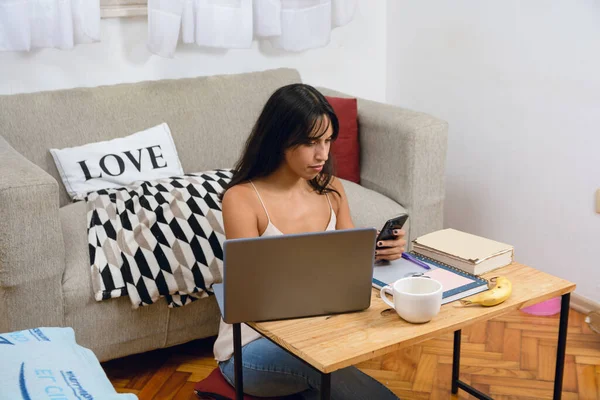 young Latina brunette entrepreneur woman at home sitting busy working with her laptop reviewing phone planning agenda and appointments with her clients.