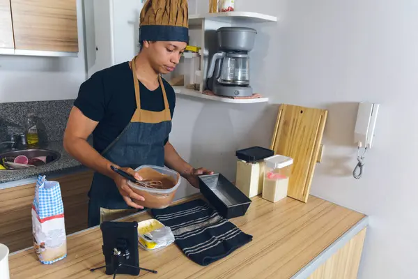 Young Latino cook man in his home kitchen, standing on counter holding container with liquid cake mix to pour into baking pan, copy space, lifestyles concept.