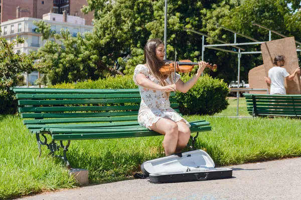 young latin woman busker violinist working sitting on wooden bench in plaza in buenos aires argentina, with case on floor for money, music and street performer concept, copy space.