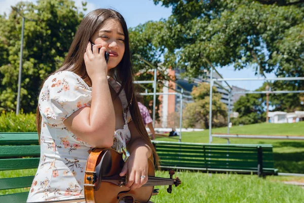 young Latin busker woman violinist with long hair and dress, is sitting with her violin in her legs, talking on phone in park at noon.