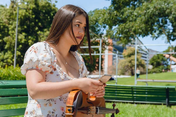 young buske latina violinist woman sitting on wooden bench outdoors, calmly checking social networks using phone, connection of people, music and technology, copy space.