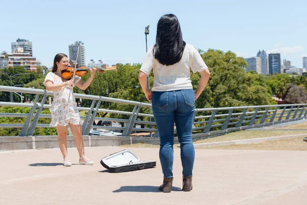 young latin woman in skirt, busker playing violin on street while another woman stands watching her and enjoys her music, music concept, copy space.