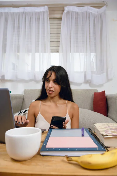 Vertical image of young Latina brunette entrepreneur at home sitting in living room using her laptop and phone, managing her business, using internet and apps.