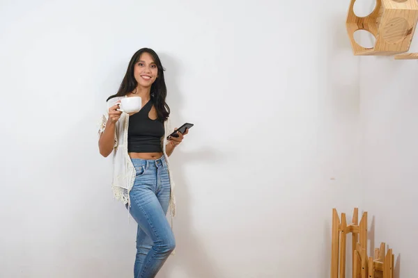 young black haired latin woman at home calmly standing using mobile looking at camera smiling with cup of coffee in morning while resting, copy space.