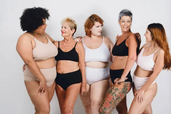 5 women of different ethnicities and ages posing, afro, caucasian, brunette, fat and thin, with short and long hair, are in underwear on white background looking at each other