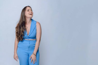 Uzun kahverengi saçlı ve mavi elbiseli genç Latin kadın, girişimci duruşu kahkaha atma, rahatlama, beyaz arka planlı stüdyo çekimi, kopyalama alanı