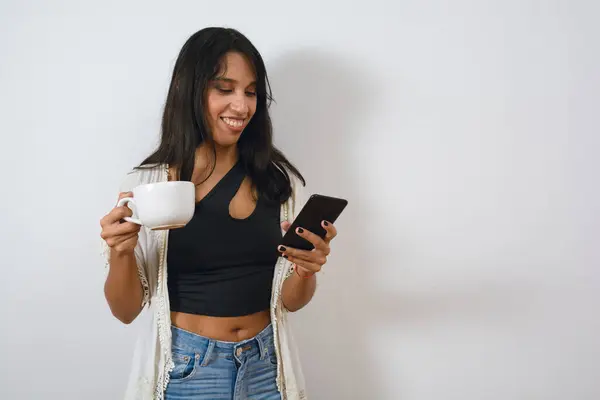 young latin woman at home standing drinking coffee and laughing watching videos on phone, browsing internet, checking social media, white wall background with copy space