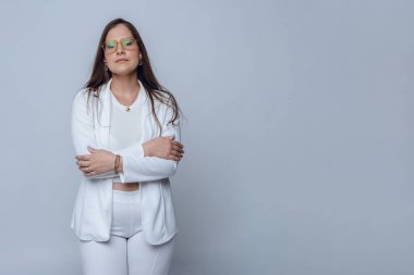 Beyaz elbiseli, gözlüklü ve uzun saçlı genç Latin iş kadınının belden yukarı portresi, kameraya bakarak kollarını kavuşturuyor. Boşluğu kopyala, beyaz arkaplan.