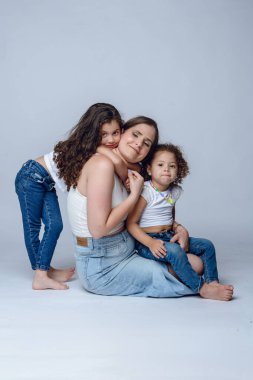 Genç Latin anne gülümseyip poz veriyor ve kıvırcık saçlı, beyaz arka planda iki güzel kızıyla poz veriyor..