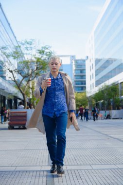 Ticari binaların önünde yürüyen, kahve içen ve etrafa bakınan girişimci genç adamın dikey görüntüsü..