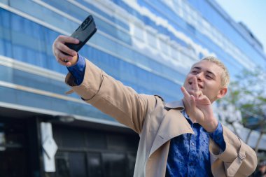 Arjantinli genç bir adam, resmi giyinmiş yakışıklı sarışın girişimci arka planda ticari binayla dışarıda telefonuyla selfie çekiyor. Boşluğu kopyala.