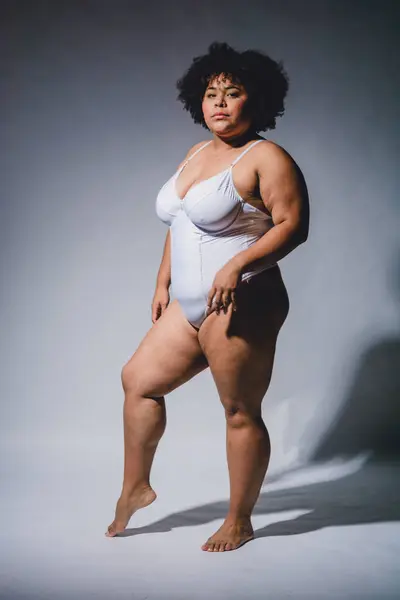 Genç Venezuelalı Latin Afro kadın, beyaz iç çamaşırlarıyla poz veriyor, kendinden emin ve güçlü, kameraya ciddi ciddi bakıyor, stüdyo fotoğrafında yüksek kontrastlı ışık var. dikey resim.