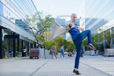 Kısa sarı saçlı genç eşcinsel adam, profilde sokak atlaması, bacaklarını kaldırması ve elleriyle ceketini sallaması, kameraya bakması, yaşam tarzı konsepti, kopyalama alanı..