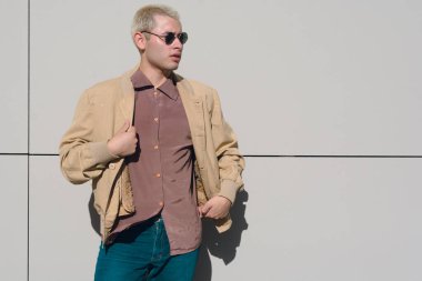 Gün batımında güneş gözlükleriyle dışarıda dikilen kahverengi ceketli genç latin adam etrafa bakıyor. Boşluğu kopyala.