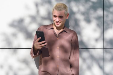 Sarı saçlı genç bir latin adam gülümsüyor ve cep telefonu tutuyor. Bir duvarın önünde duruyor.