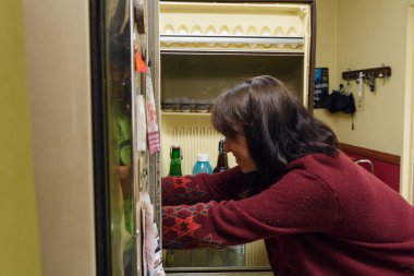 Yetişkin bir Latin kadın buzdolabına yemek bakıyor ve gülümsüyor. Kırmızı bir kazak giyiyor ve elinde bir şişe bira var. Yaşam tarzı konsepti, fotokopi alanı..