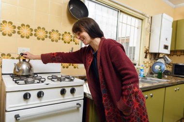 Arjantinli yetişkin kadın mutfakta su ısıtmak, kahvaltı yapmak ve güne başlamak için ocağa su koyuyor, kırmızı kazak giyiyor ve gülümsüyor.