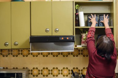 Mutfakta elleri havada duran, dolaptan kahve kutusunu alan, kırmızı kazak giyen, yeşil martılı klasik bir ev ortamında olan tanınmamış bir kadının arka görüntüsü.