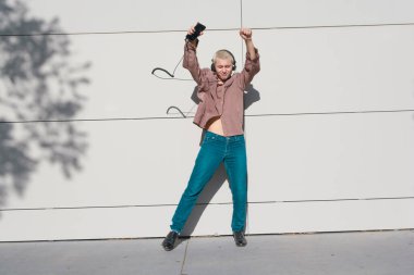 Gün batımında kısa saçlı, kahverengi gömlekli ve kot pantolonlu, kulaklıklı dans eden ve müzik dinleyen genç sarışın adam günbatımının tadını çıkarıyor..