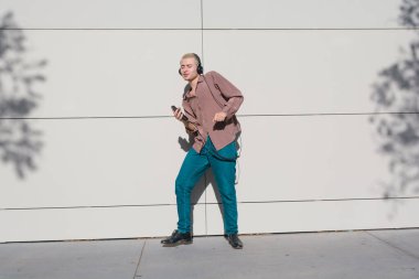 Gün batımında kısa saçlı, kahverengi gömlekli ve kot pantolonlu, kulaklıklı dans eden ve müzik dinleyen genç sarışın adam günbatımının tadını çıkarıyor..