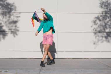 Kısa sarı saçlı, pembe şortlu, siyah spor ayakkabılı ve yeşil kazaklı genç Latin adam, açık havada gururlu bir şekilde dans ediyor ve günbatımının tadını çıkarıyor..