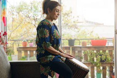 Kanepede oturan bir kadın davul çalıyor. Sahne huzurlu ve rahatlatıcı. Kadın enstrüman çalmaktan zevk alıyor.